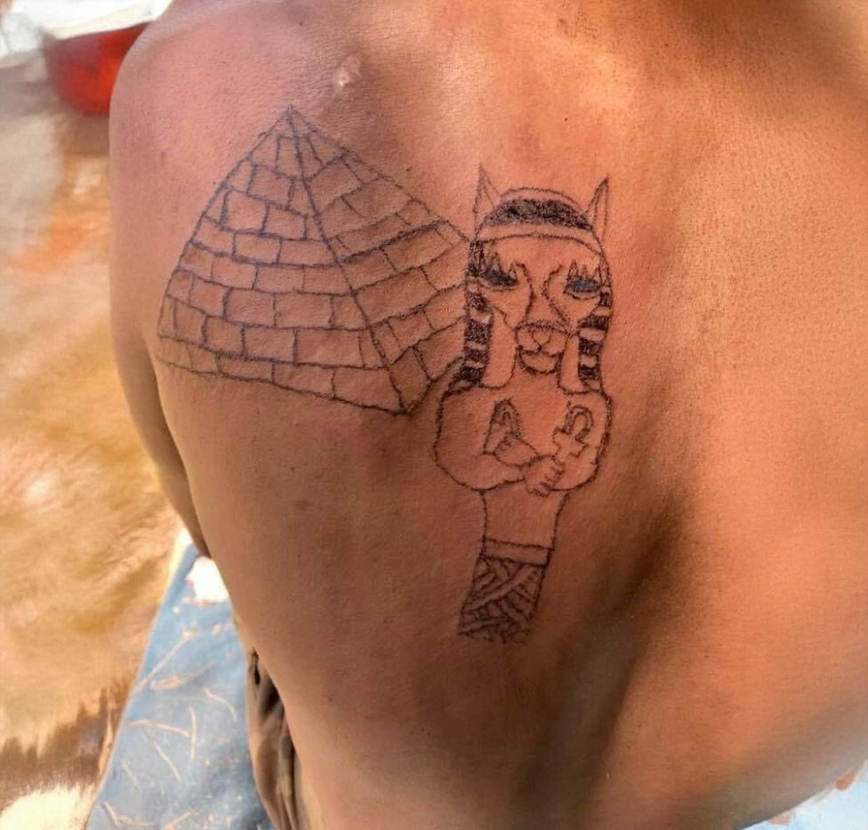 A person’s back with a tattoo of a pyramid and a poorly drawn ancient Egyptian pharaoh or sphinx figure with crossed arms and an exaggerated face. The tattoo appears to be done in black ink.