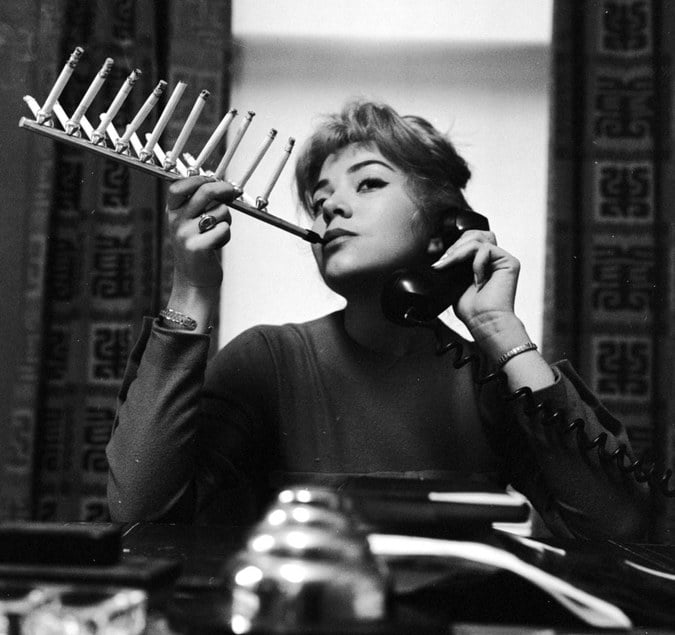 A woman sits at a desk holding a telephone receiver to her ear while using a large cigarette holder to smoke multiple cigarettes at once. She looks upward thoughtfully.