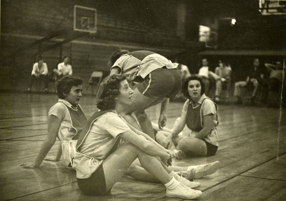 Four women in athletic uniforms sit and kneel on a gymnasium basketball court, appearing relaxed during a break. The background shows empty seats, a basketball hoop, and a few people on the sidelines.