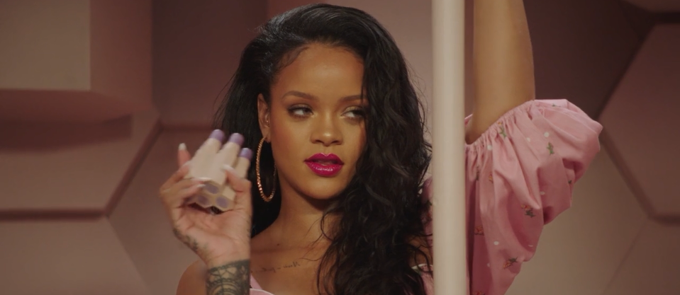 A woman with long dark hair in a pink off-shoulder dress holds a makeup product and poses with one arm raised, standing against a pink geometric background. She has bright pink lipstick and wears hoop earrings.