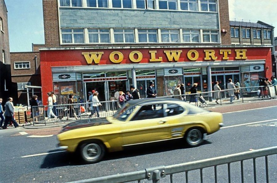 A yellow classic car drives past a Woolworth store with a bright red facade, while people walk along the sidewalk in front of the building on a sunny day.