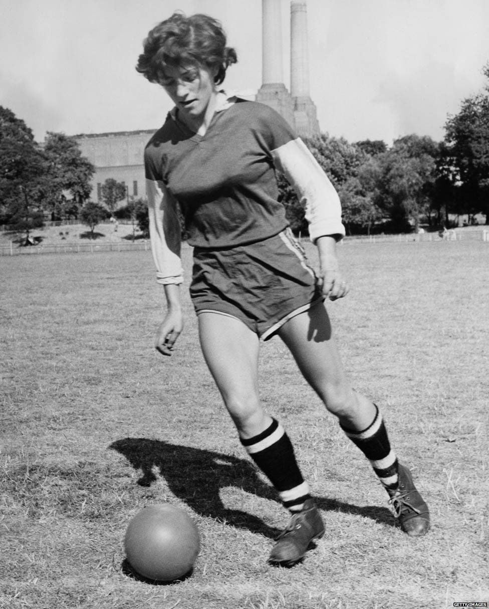 A woman in a soccer uniform runs on a grassy field while dribbling a soccer ball. Buildings and trees are visible in the background under a clear sky.