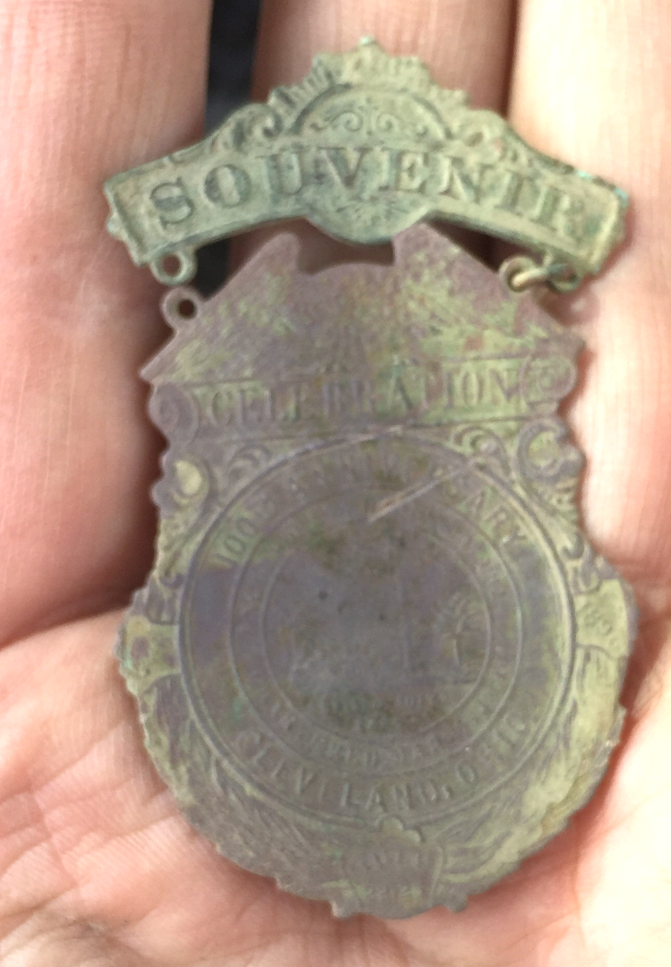 A person's hand holds a worn, tarnished metallic souvenir badge with engraved text and designs; "SOUVENIR" is visible at the top, but most other details are faded and difficult to read.