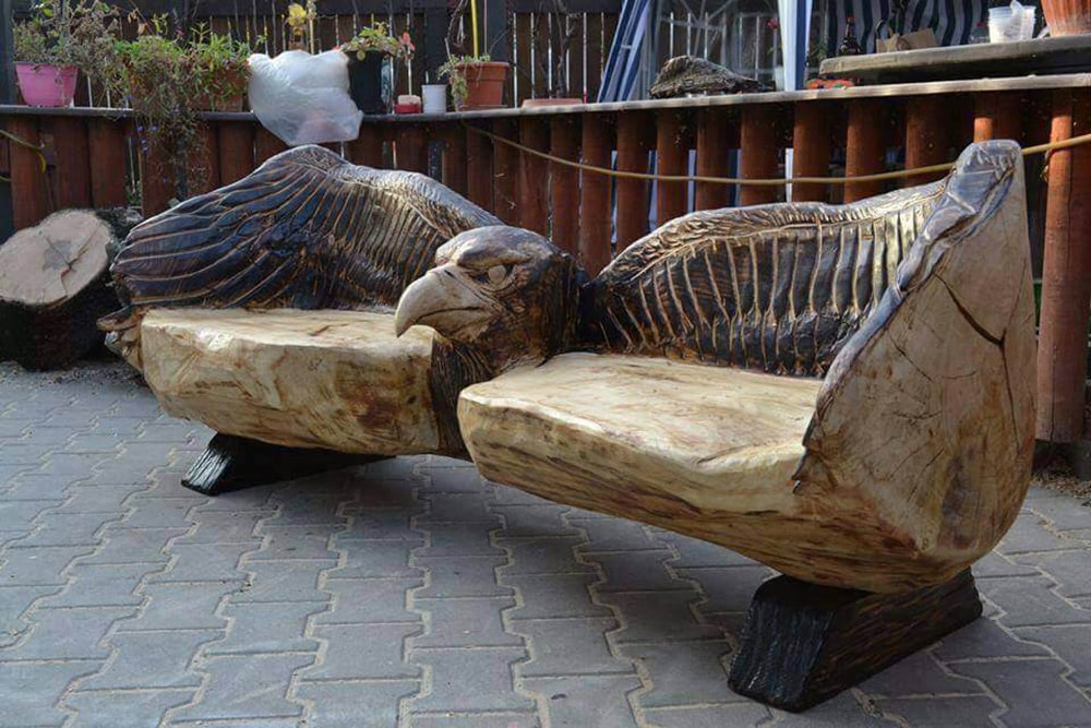 A wooden bench intricately carved to resemble a large eagle with outstretched wings, forming the seat and backrest. The bench is placed on a stone-paved outdoor area.