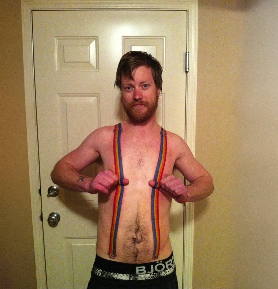 A man with a beard stands indoors in front of a door, wearing only underwear. He has rainbow stripes painted vertically over his chest and follows the lines with his thumbs.