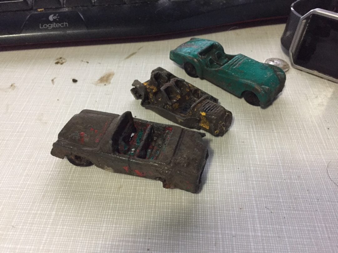 Three small, weathered toy cars are placed on a light-colored surface, with a keyboard and digital watch in the background. The toy cars appear old and worn, with faded paint and missing wheels.