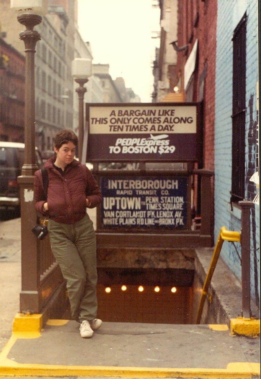 A person with short hair, wearing a maroon sweater and green pants, stands at the entrance of a subway station in a city. Signs above advertise bus fares to Boston and subway routes. Urban buildings are visible in the background.