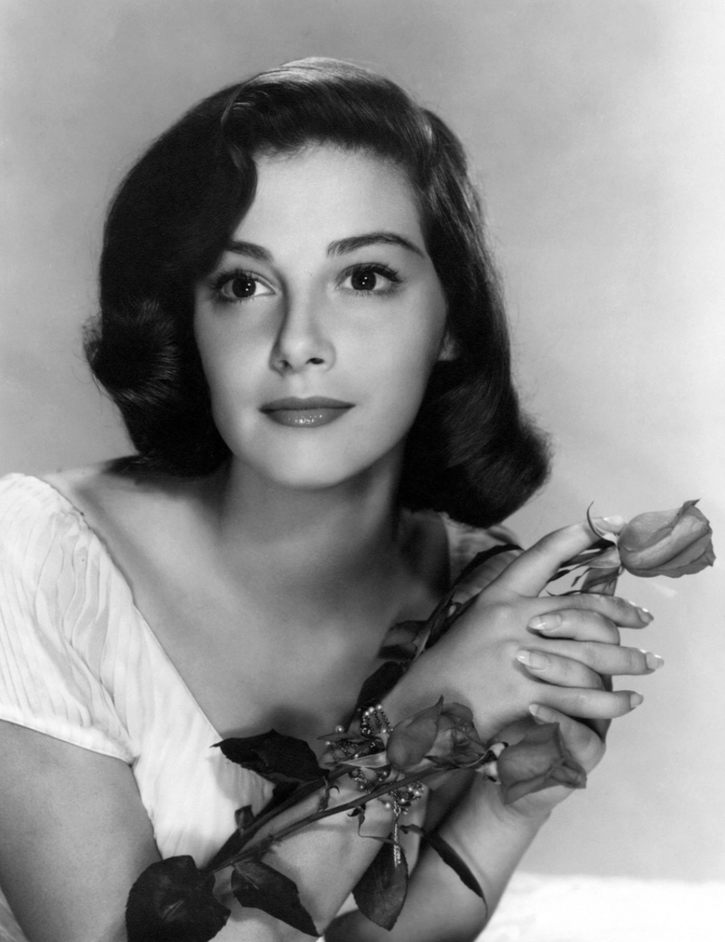 Black and white portrait of a young woman with short, wavy dark hair, wearing a light dress, holding a rose with both hands, and gazing slightly upward with a serene expression.