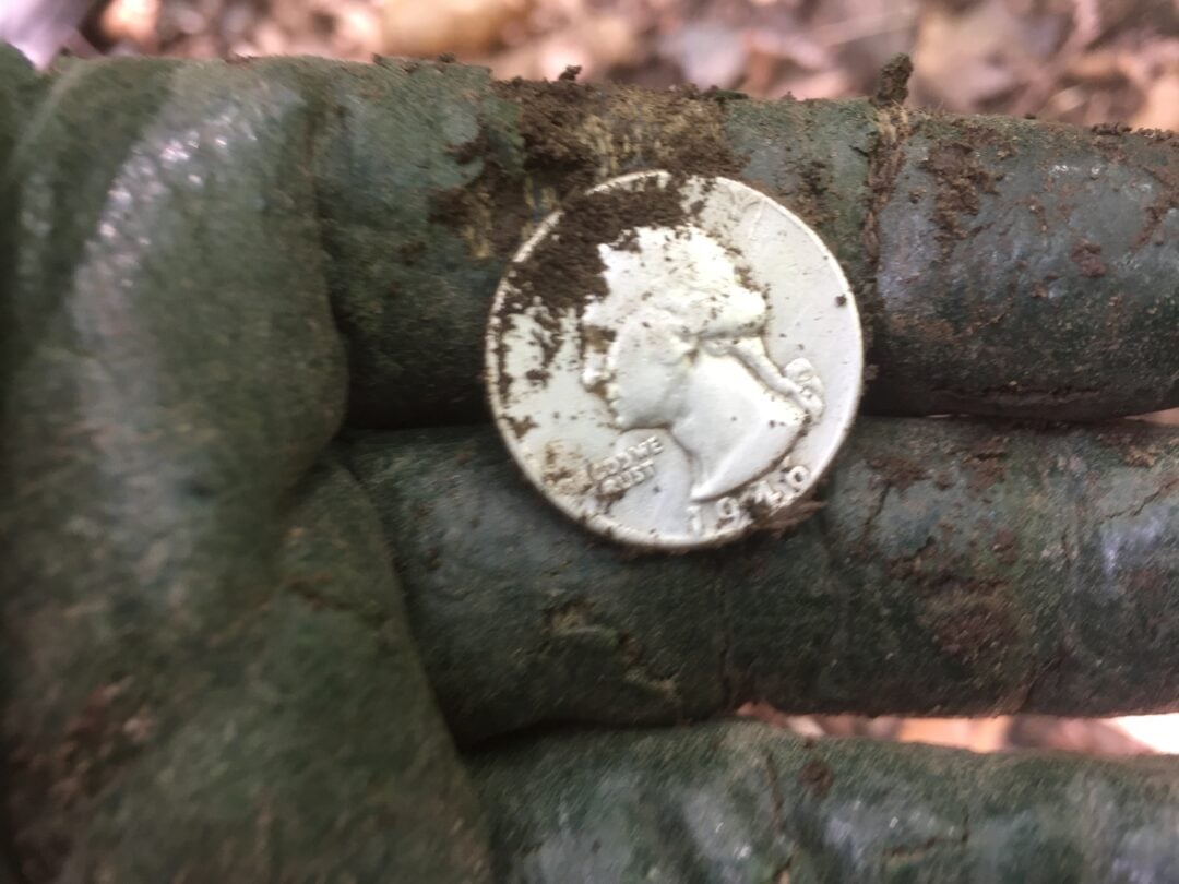 A muddy gloved hand holds a dirty quarter coin with the profile of a woman visible, partially covered in soil. The background shows blurred earth and leaves.