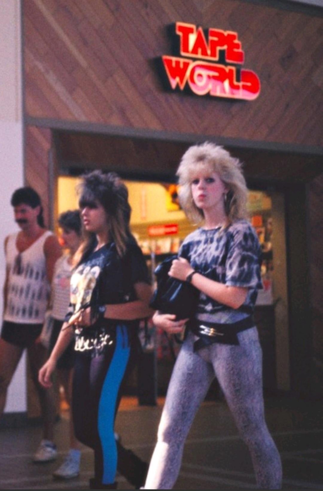 Two women with teased, 1980s-style hair and bold clothing walk past a store called "Tape World" in a mall. Several other people are in the background, also dressed in casual '80s attire.