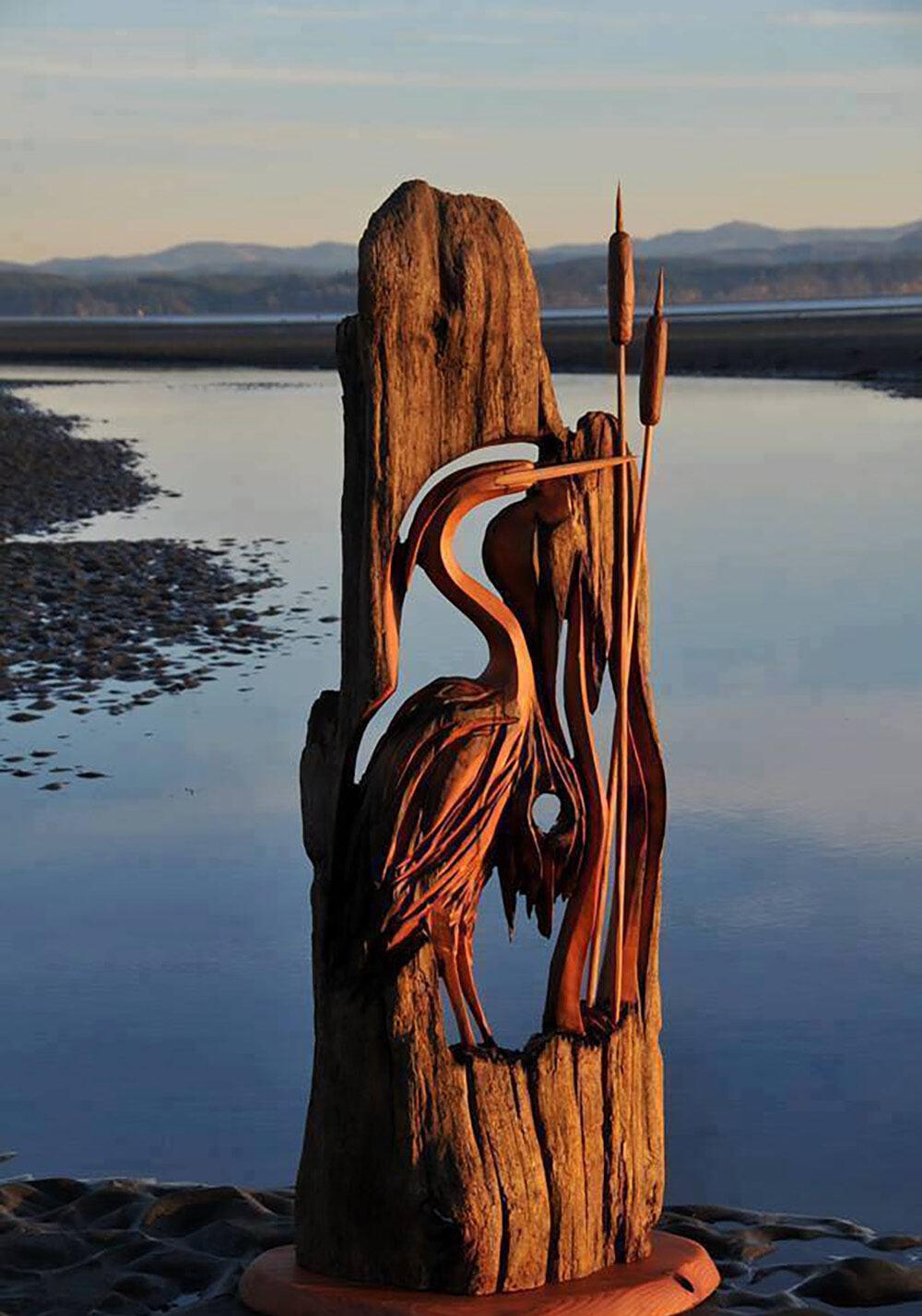 A wooden sculpture of a heron and cattails carved into a tree trunk stands on the shore of a calm body of water at sunset, with distant mountains in the background.