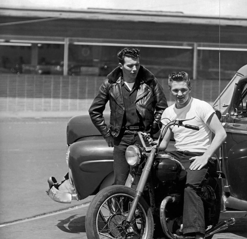 Two young men in 1950s-style clothing hang out beside an old car; one in a leather jacket stands while the other, in a white T-shirt, sits on a classic motorcycle, both appearing relaxed.