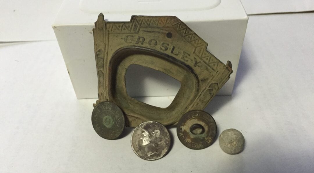 A vintage metallic frame labeled “Crosley” is displayed upright, with three old coins or tokens and a small round object arranged in front of it on a white surface.