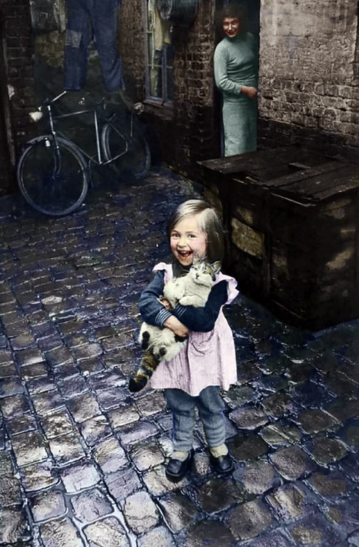 A smiling young girl stands on a wet cobblestone street, holding a cat in her arms. She wears a pink dress over clothes. Behind her, a woman in a green dress looks out from a doorway. A bicycle leans against the wall.