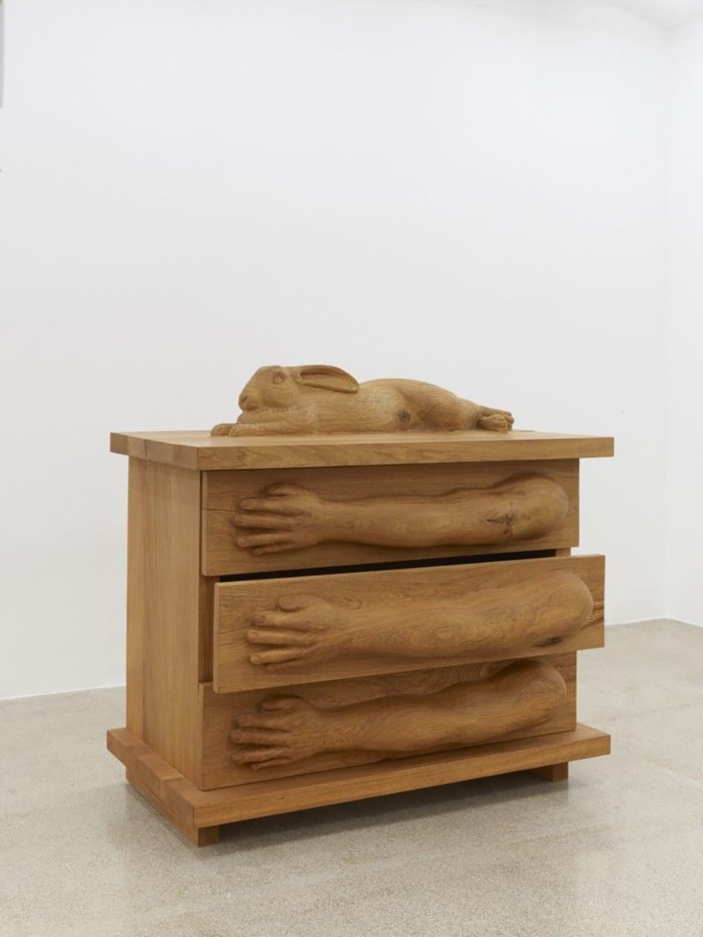 A wooden chest of drawers with sculpted human arms as drawer handles and a carved rabbit lying on top, set in a plain white room.