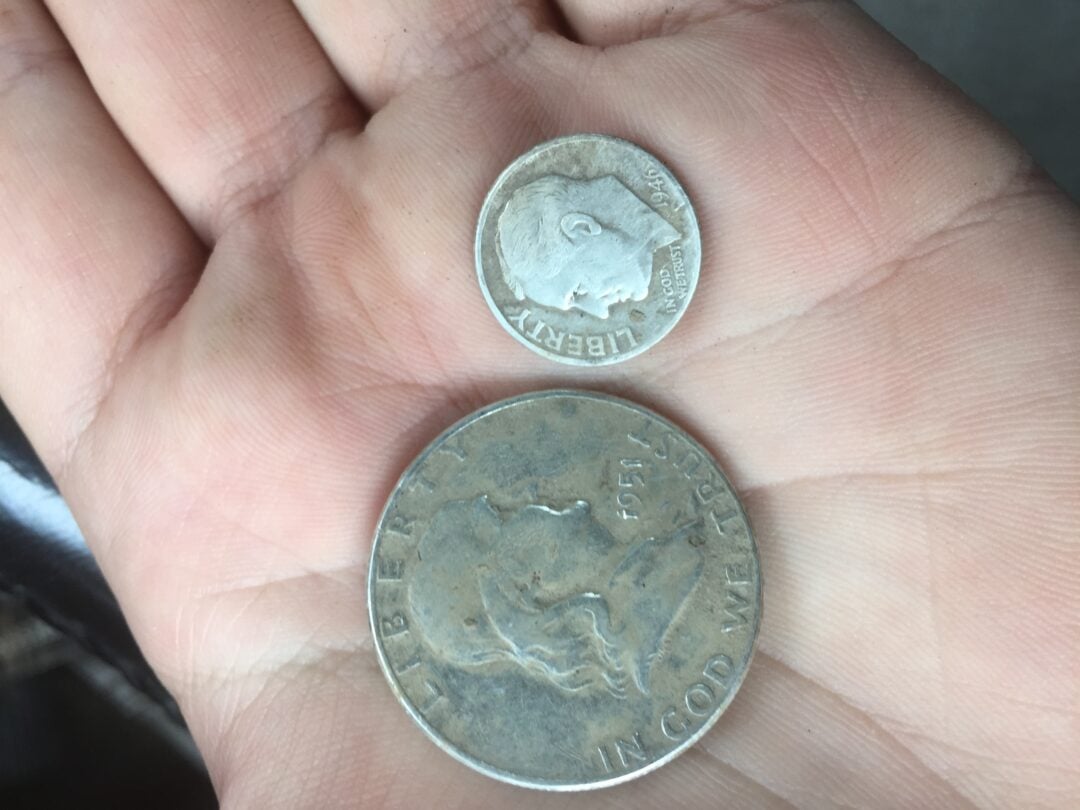 A hand holding two U.S. coins: a small silver dime above a larger silver half dollar. Both coins display profiles of different people and the word "LIBERTY" is visible on each.