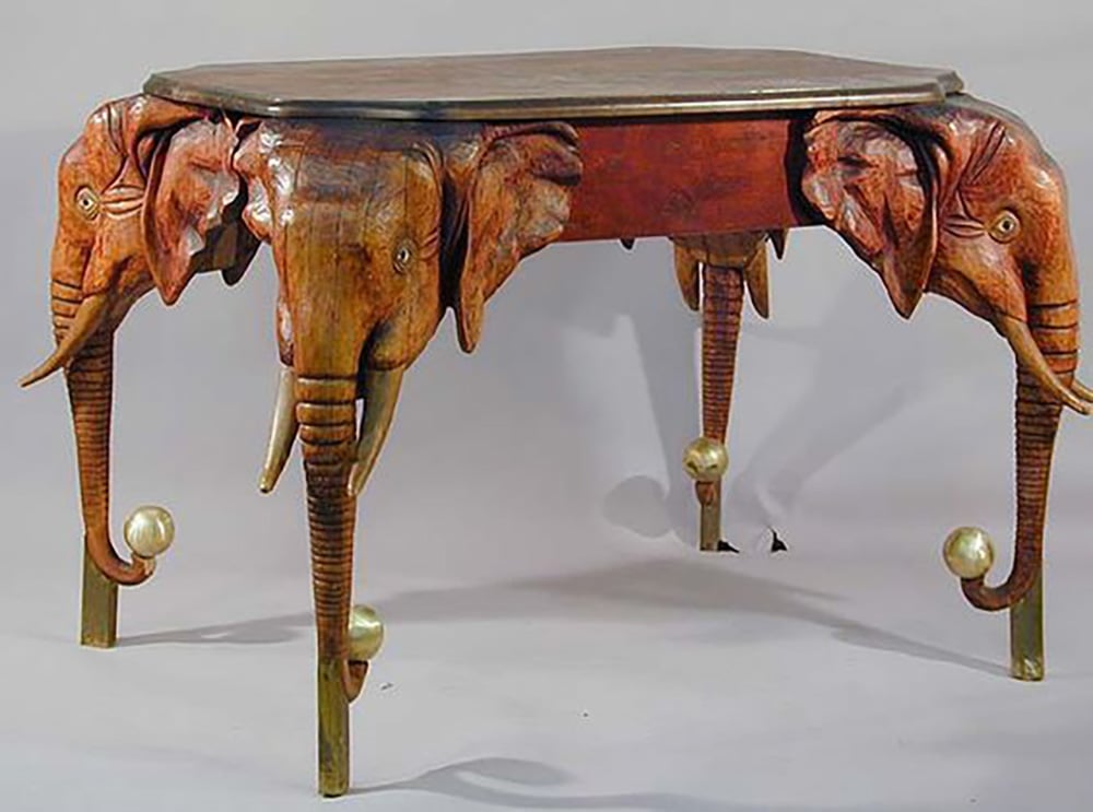 A wooden table with three legs, each carved to resemble an elephant’s head and trunk. The trunks curve downward to form the table legs, with gold-colored balls at the end of each trunk.