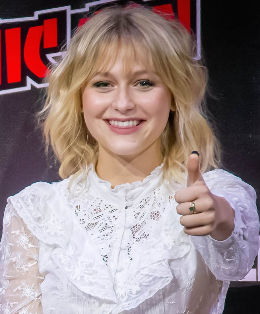 A woman with blonde, wavy hair and bangs, dressed in a white lacy blouse, smiles warmly and gives a thumbs-up gesture. She is standing in front of a dark background with red and white text.