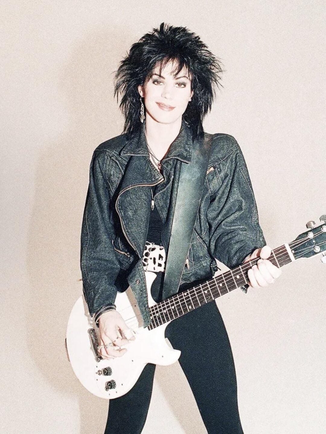 A person with spiky black hair, wearing a black denim jacket, black pants, and a studded belt, smiles while holding a white electric guitar against a plain light background.