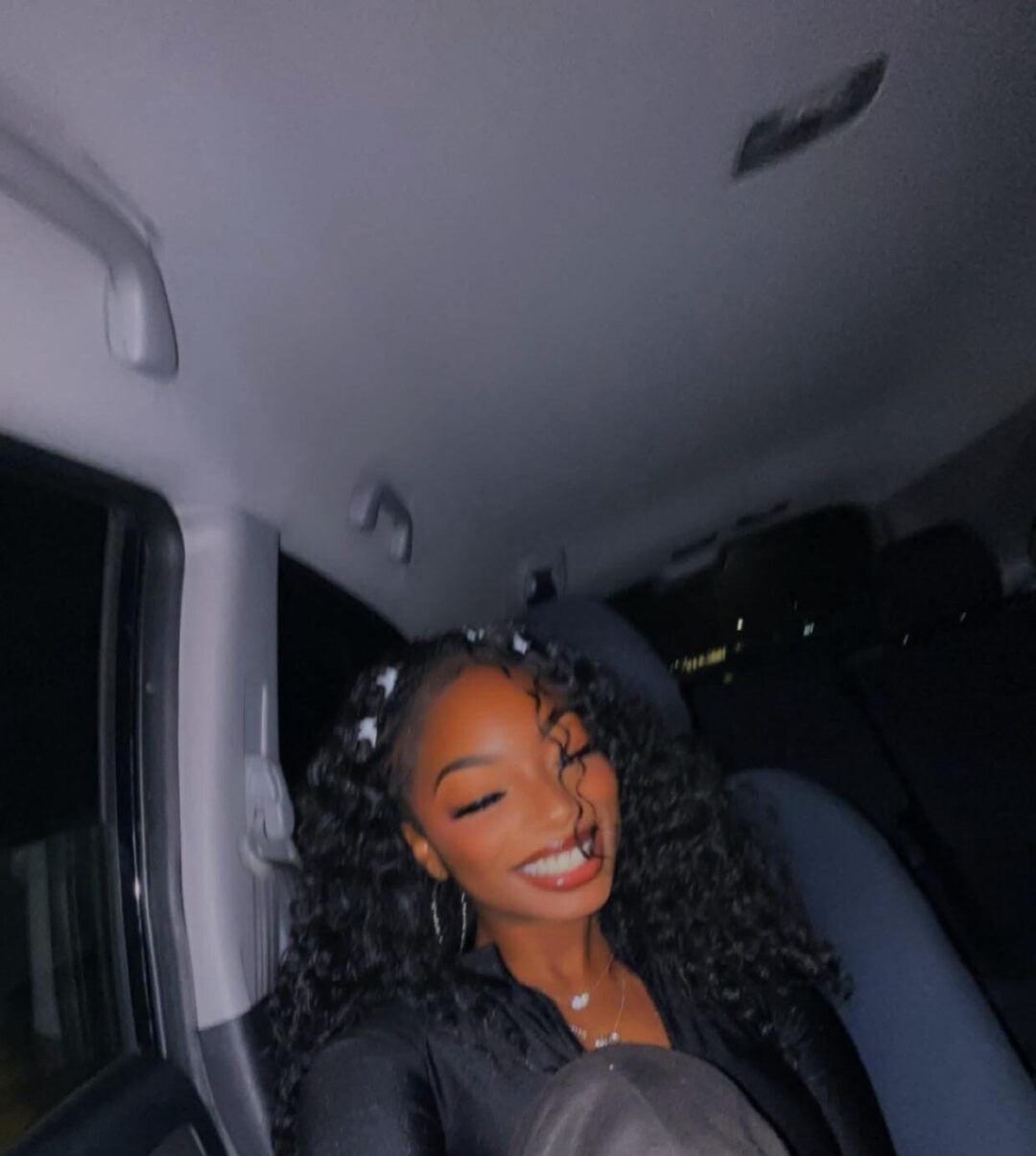A woman with long, curly hair sits in the backseat of a car, smiling brightly at the camera. She is wearing a black top and hoop earrings, with one knee up in front of her. The photo is taken at night.