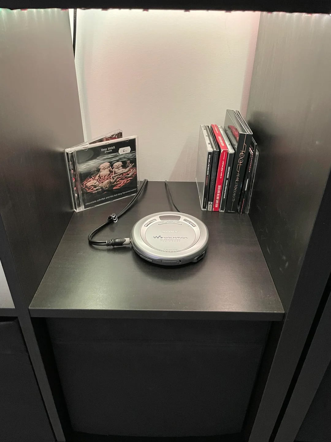 A silver Sony Discman CD player with attached earbuds sits on a black shelf. Behind it are several CD cases, including one with visible album art and others stacked vertically.