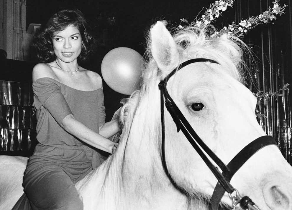 A woman in a red dress sits on a white horse indoors, smiling. A man in a red jacket and bow tie leads the horse. Several people stand nearby, watching and smiling. The scene is festive, with balloons on the floor.