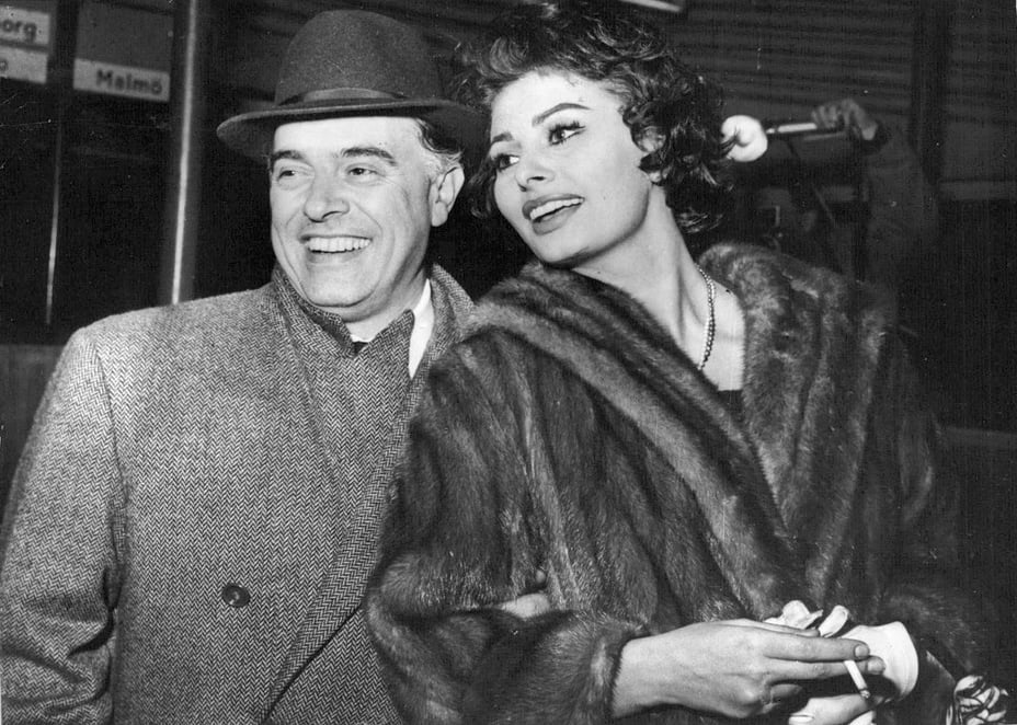 A man in a hat and coat stands smiling beside a woman wearing a fur coat. The woman is looking away, holding gloves and a purse. They appear to be at a public event or gathering.