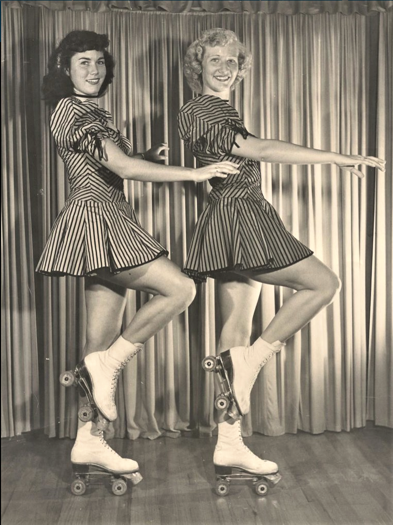 Two women in matching striped dresses and roller skates pose side by side on one leg each, arms raised slightly, in front of a curtain backdrop. The image is black and white and appears to be vintage.