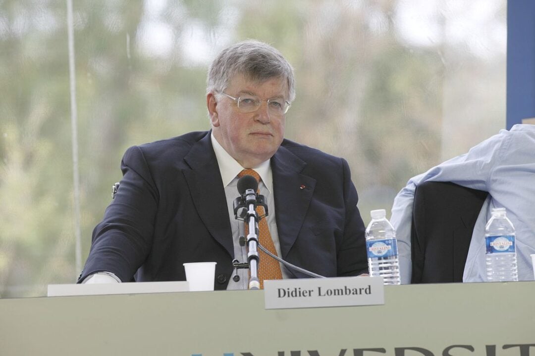 A man in a suit and tie sits at a table with a microphone and water bottles in front of him. A nameplate reading "Didier Lombard" is visible on the table. Trees can be seen in the blurred background.