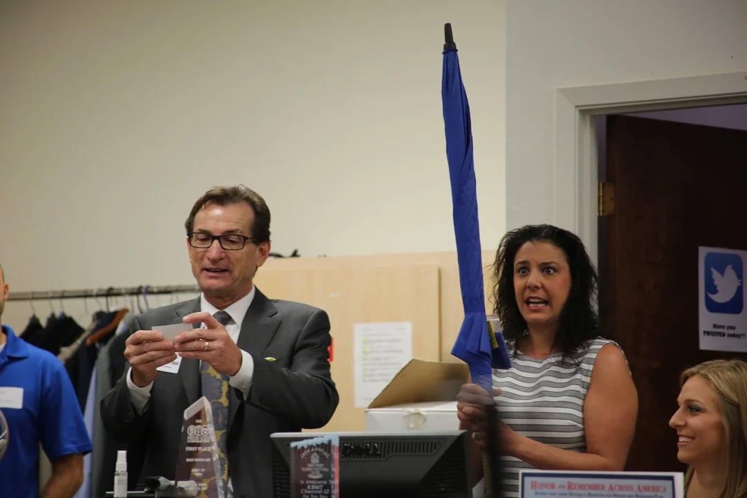 A man in a suit reads from a card while a woman beside him excitedly holds up a closed blue umbrella. Other people are partially visible around them in what appears to be an office or event space.