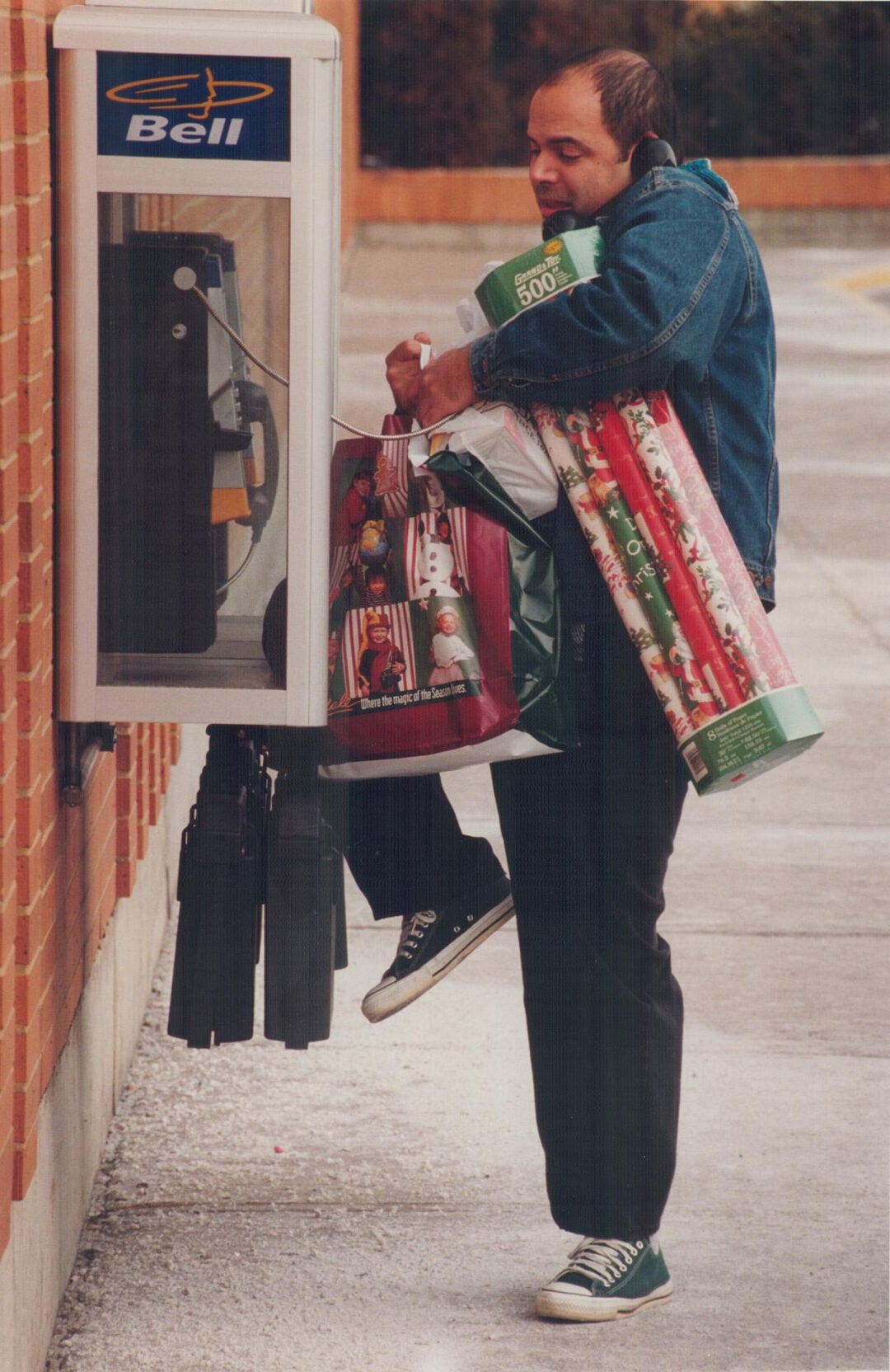 A man in a denim jacket, carrying multiple shopping bags and rolls of wrapping paper, uses his foot to open a public phone booth while balancing his items on a sidewalk.