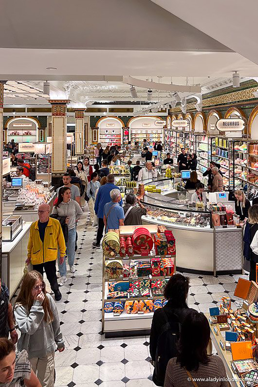 A busy, elegant food hall with tiled floors, ornate decor, and many people browsing counters and displays filled with food, drinks, and gift items. Bright lighting and high ceilings add to the lively atmosphere.