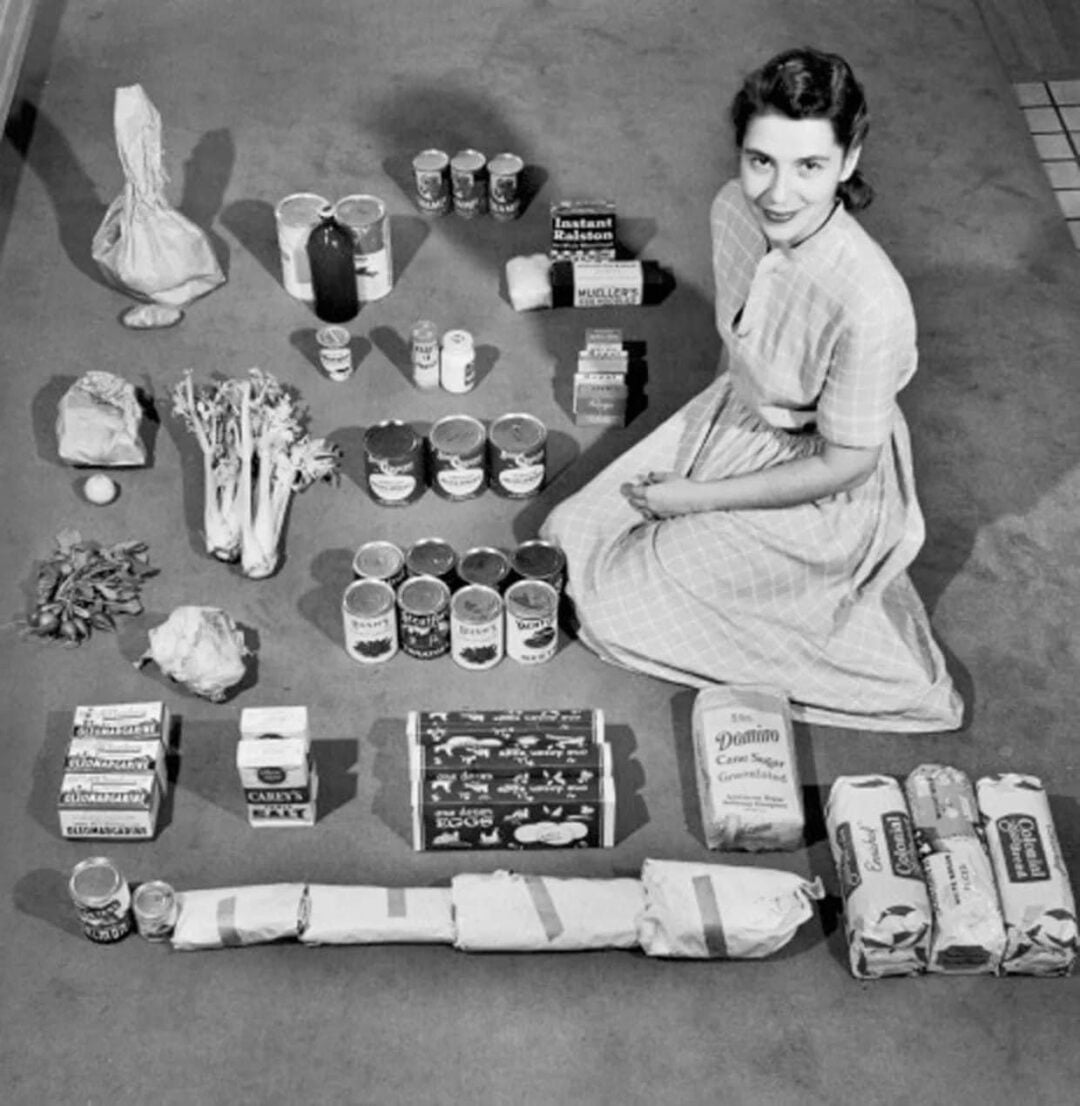 A woman in a striped dress sits on the floor next to an organized display of groceries, including canned goods, produce, sugar, flour, spices, and packaged items, all neatly arranged in rows.