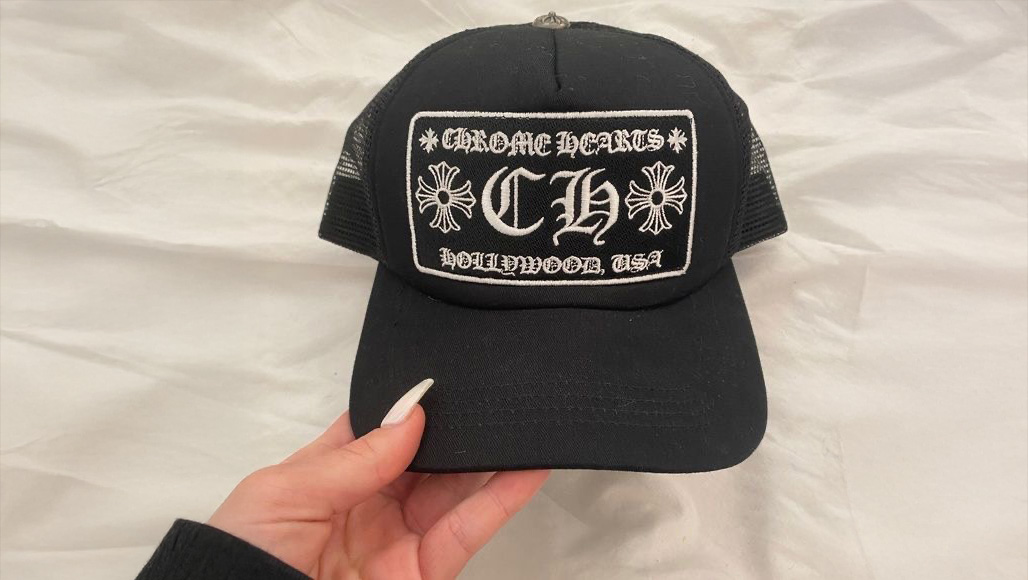 A hand holds a black trucker hat with a white embroidered patch that reads "CHROME HEARTS CH HOLLYWOOD, USA" and features two cross designs on either side of the text.