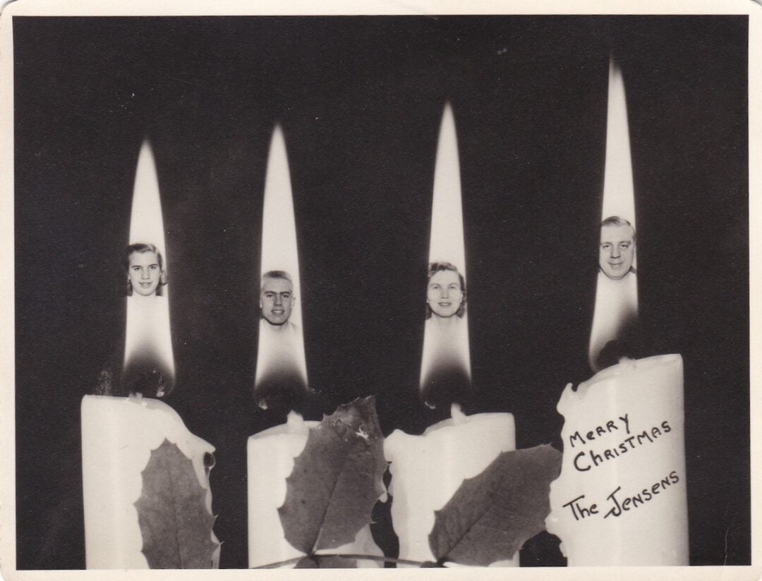 Black-and-white photo of four lit candles, each with a cut-out photo of a face in its flame. Holly leaves lay at the base of the candles. The words “Merry Christmas The Jensens” are written on one candle.