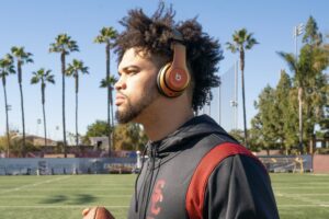 A young man wearing headphones and a USC tracksuit holds a football while standing on a football field lined with palm trees under a clear blue sky.