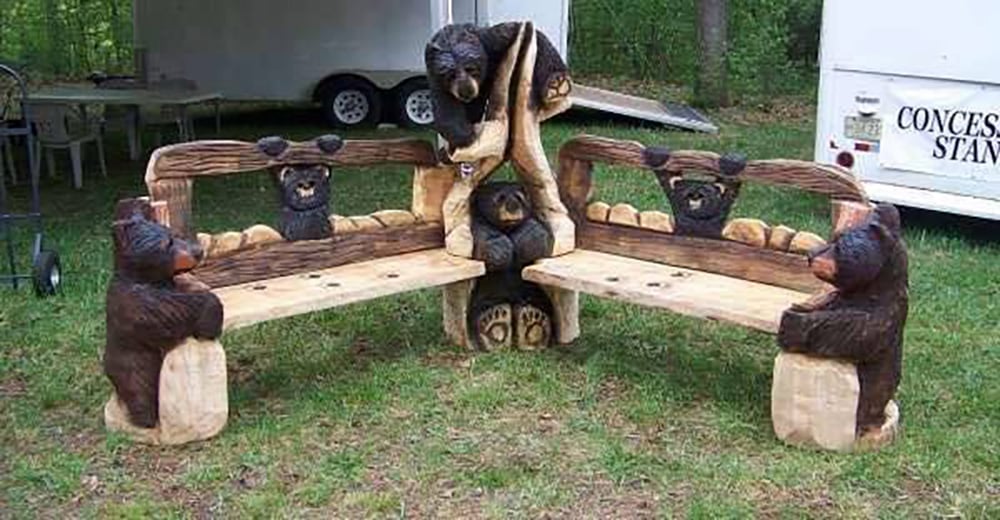 Two wooden benches feature detailed carvings of playful black bears, with additional bear carvings connecting and supporting the benches at the corners, displayed outdoors on grass near a white trailer.
