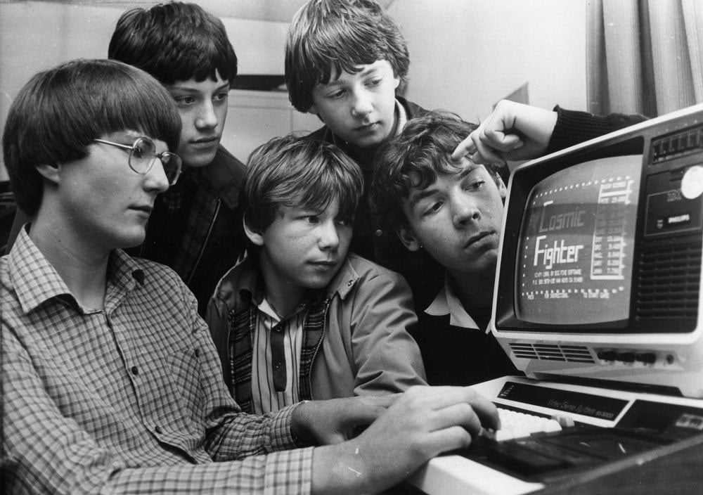 Five boys gather around an old computer, intently looking at the screen displaying a game called "Fighter." One boy points at the screen while another uses the keyboard, suggesting teamwork and curiosity.