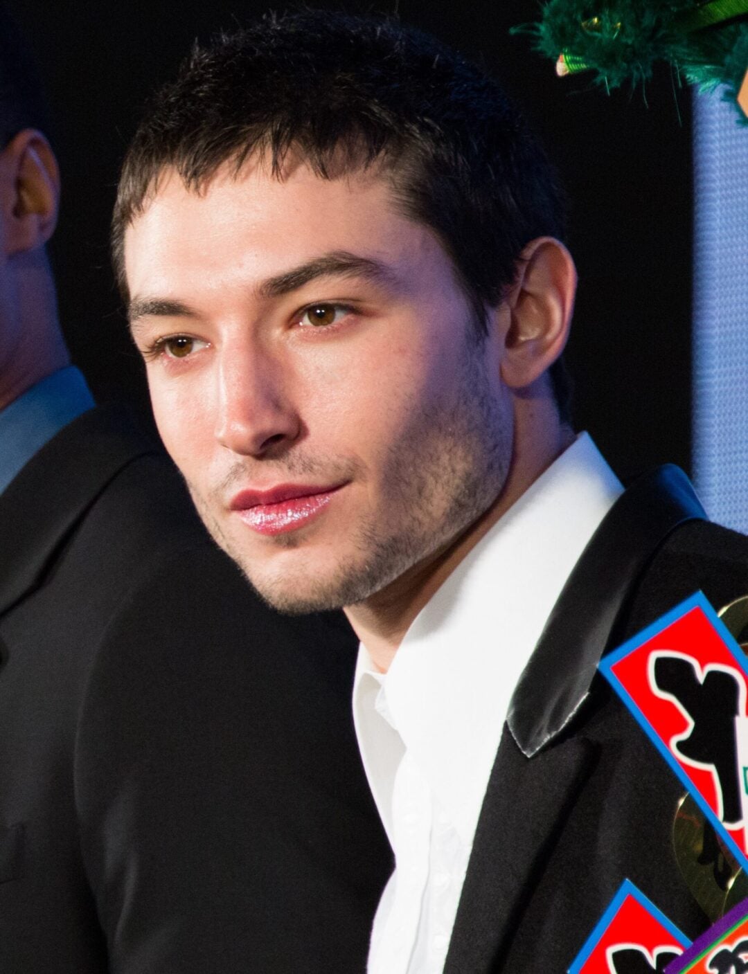 A person with short dark hair and light skin is wearing a black jacket with a white shirt. They are looking slightly to the side and standing in front of a dark background with some colorful shapes visible.