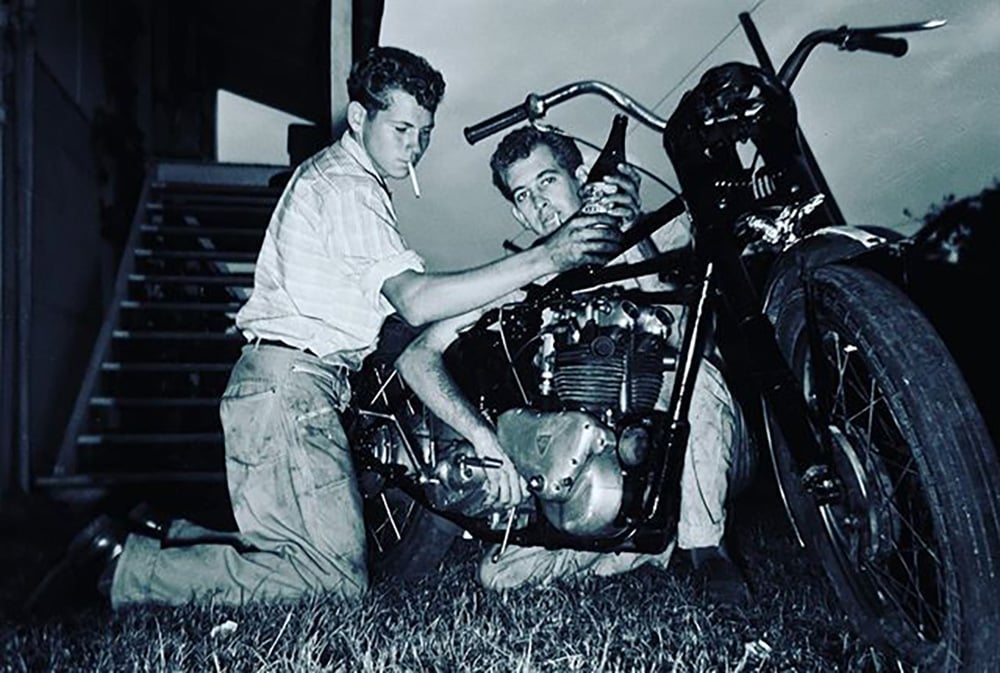 Two men kneel on grass working on a motorcycle; one holds a tool while the other adjusts the engine, and one has a cigarette in his mouth. There are stairs in the background and the scene appears to be outdoors at dusk.