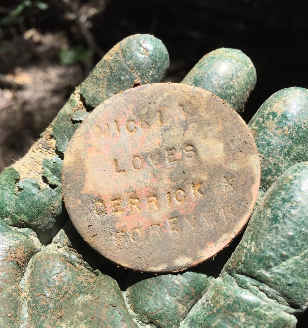 A gloved, dirt-covered hand holds a round, weathered metal token that reads, “MICHAEL LOVES JERRICK FOREVER.”