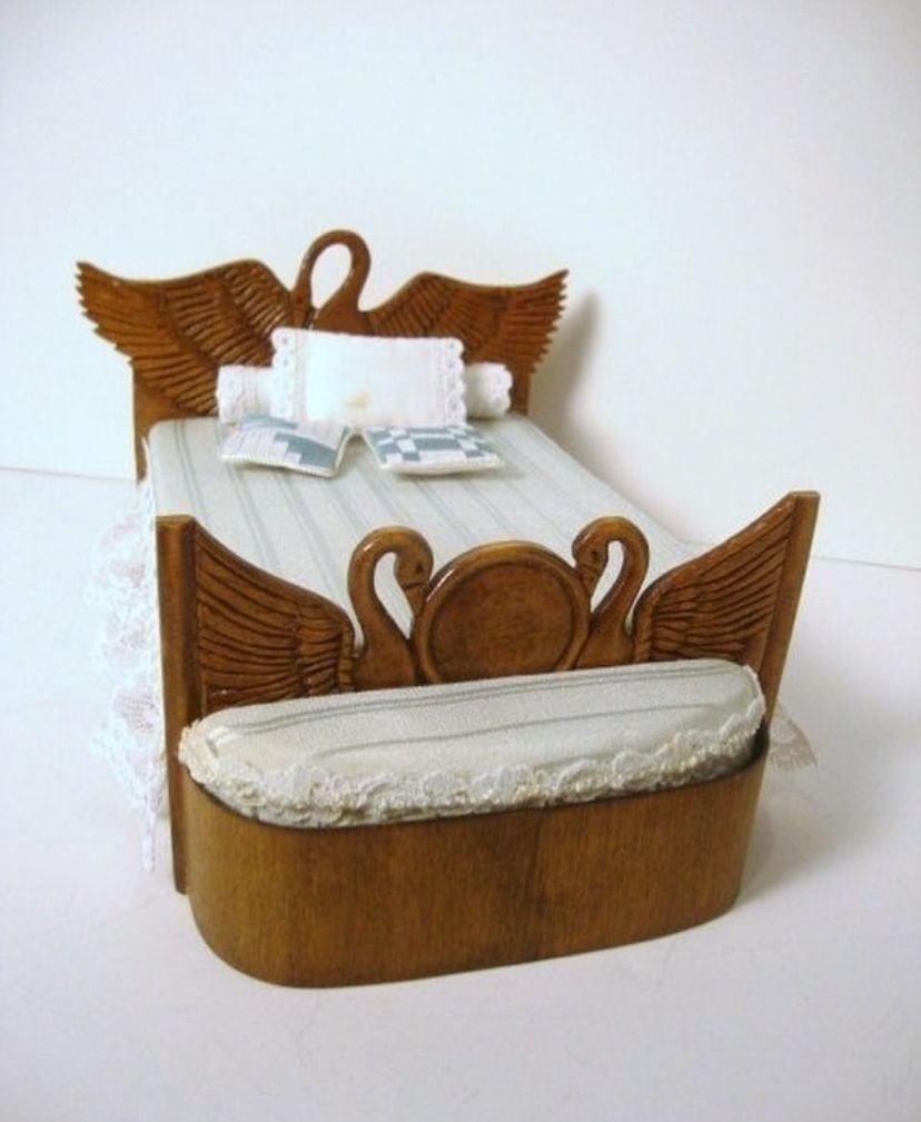 A miniature wooden bed with carved swan designs on the headboard and footboard, covered in a lace-trimmed blanket and small pillows. A matching upholstered bench sits at the foot of the bed.