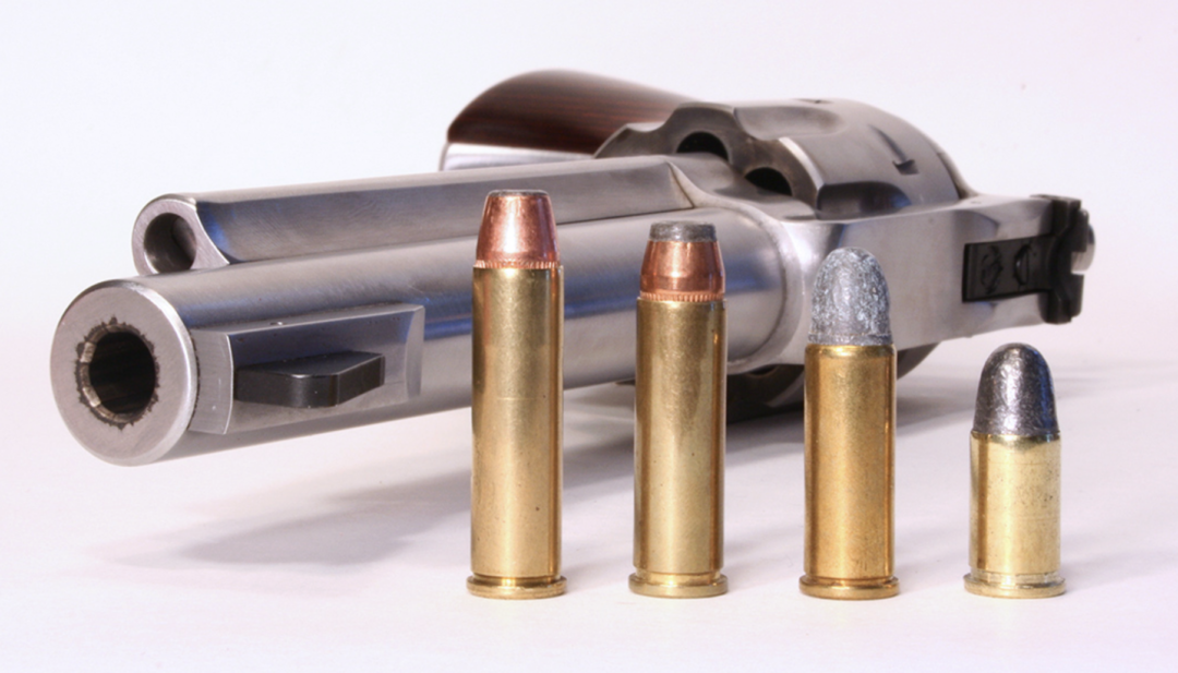 A silver revolver is lying on its side, with four different types of ammunition standing upright in front of the barrel. Each bullet has a brass casing and varies in size and tip material.