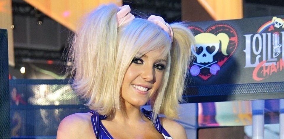 A woman in a colorful cheerleader costume with "San Romero Knights" on her top holds a toy chainsaw. She has blonde pigtails, white thigh-high socks, red leg warmers, and stands in front of a "Lollipop Chainsaw" game display.