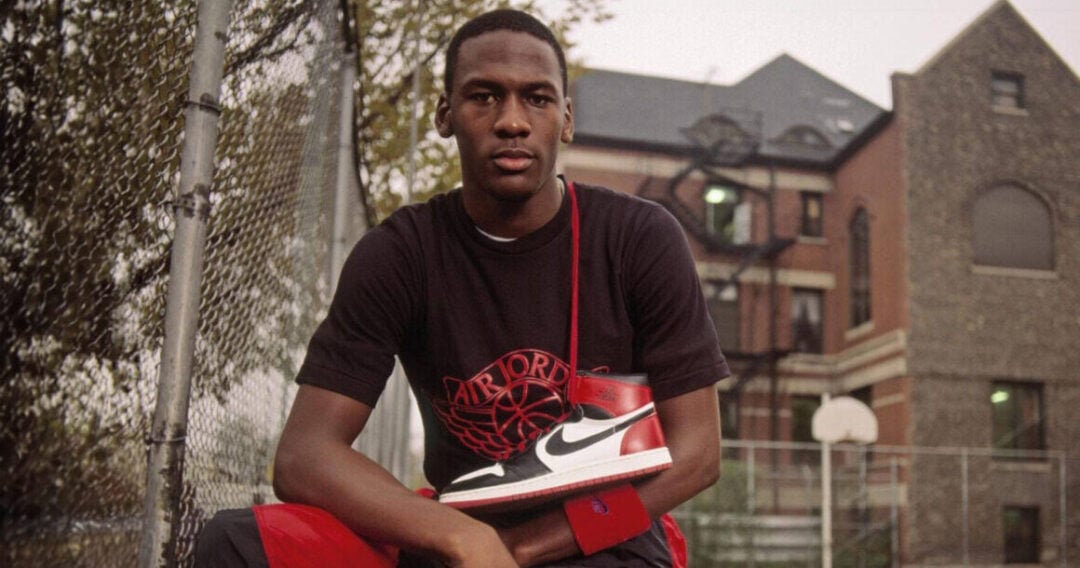 A young man sits on an outdoor basketball court, holding a red, white, and black Nike Air Jordan sneaker. He wears a black Air Jordan shirt and red shorts, with a school building and basketball hoop in the background.