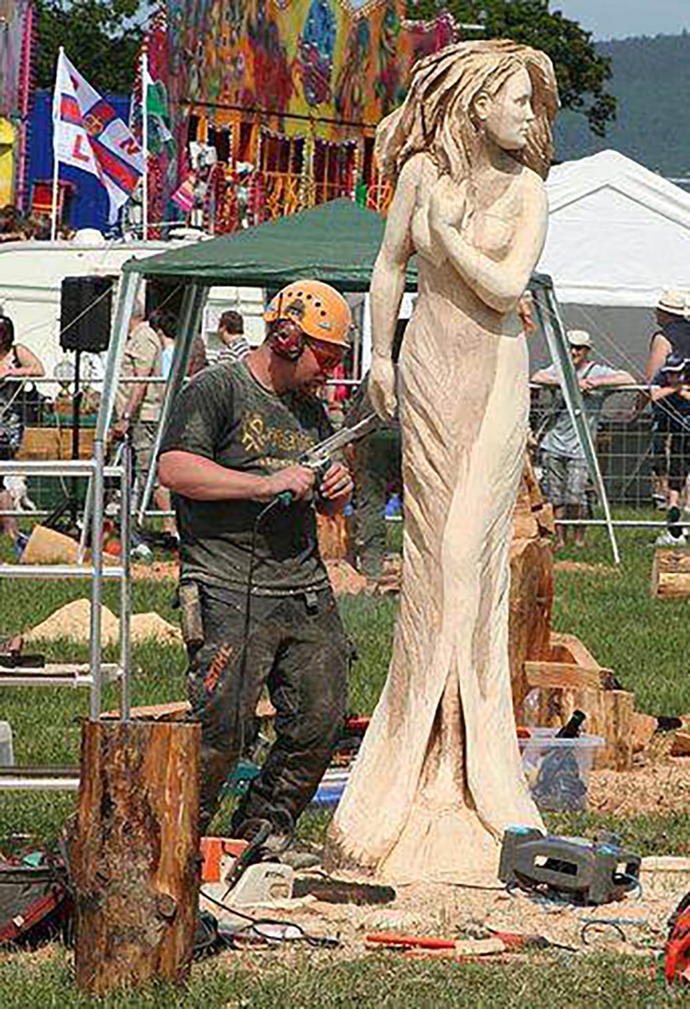 A man wearing safety gear carves a large wooden statue of a woman in a dress at an outdoor event, with tents, flags, and people in the background.