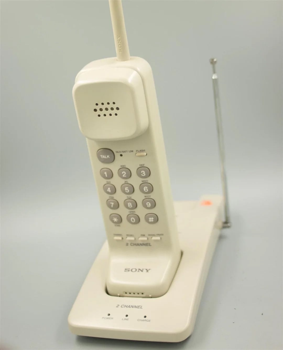 A vintage white Sony cordless landline phone with a numeric keypad, standing upright in its charging base, featuring an antenna and labeled buttons.