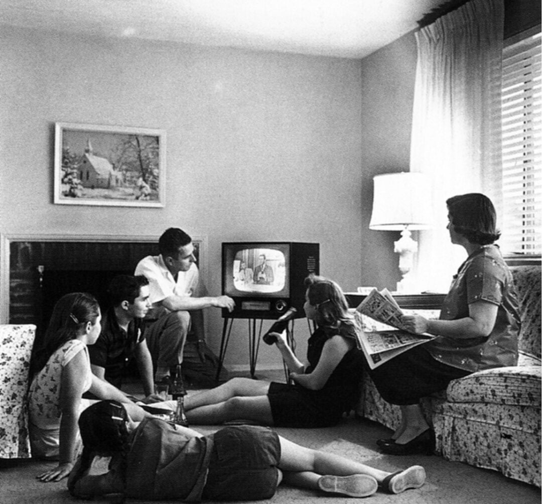 A family of five in a living room watches a black-and-white TV. Two girls lie on the floor, two adults and one child sit on sofas. The mother holds a newspaper. Sunlight streams in through window blinds.