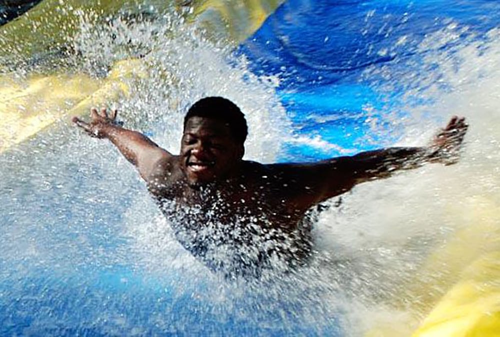 A person slides down a water slide with arms outstretched, smiling joyfully as water splashes around them on a blue and yellow slide.