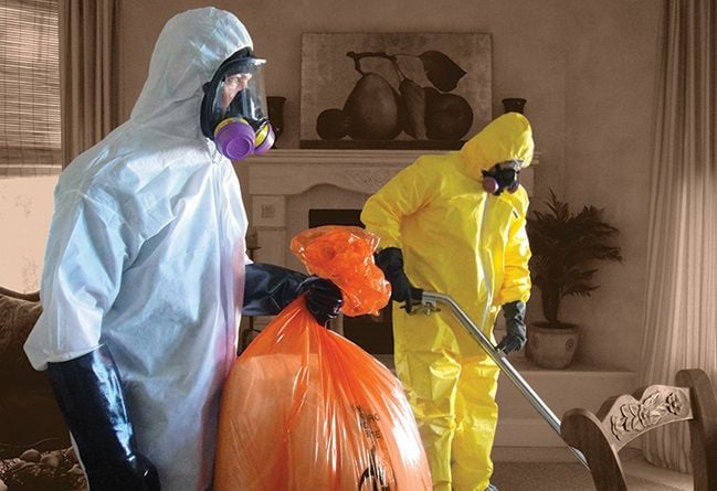 Two people in hazmat suits and respirators clean a living room; one holds an orange bag while the other uses cleaning equipment. The room is neutral-toned with curtains, a chair, and artwork visible.